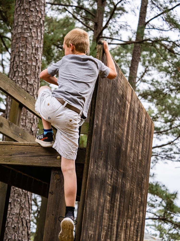 A boy in a gray shirt climbs up a wooden structure