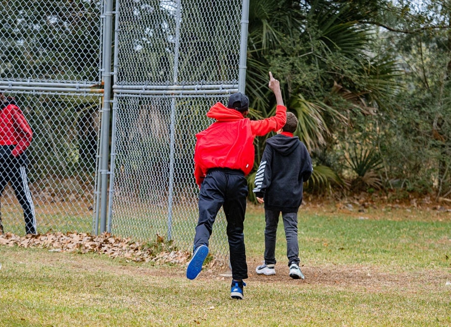 A boy in red gestures to his kickball team near the batting fence
